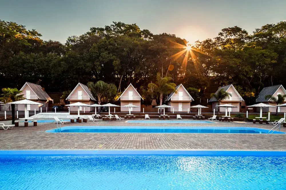 Fileira de cabanas em estilo chalé atrás da piscina, com o sol se pondo entre as árvores
