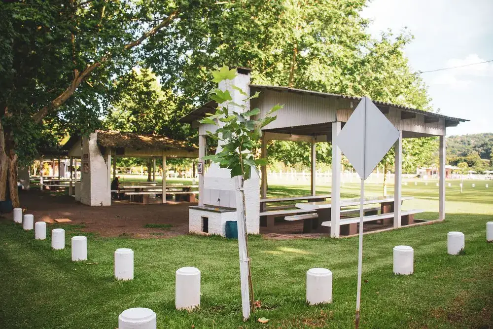 Quiosque coberto com mesas e churrasqueira, no gramado entre as árvores