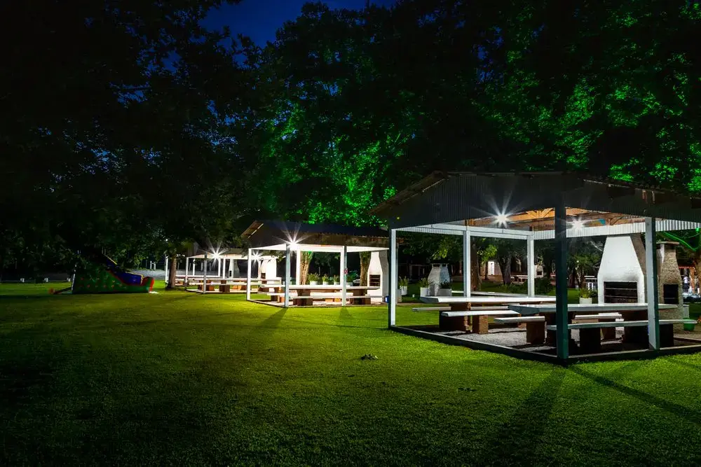 Quiosques cobertos com mesas e churrasqueira, iluminados à noite no gramado